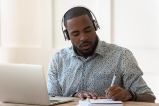 Person in front of the laptop with headphones and taking notes
